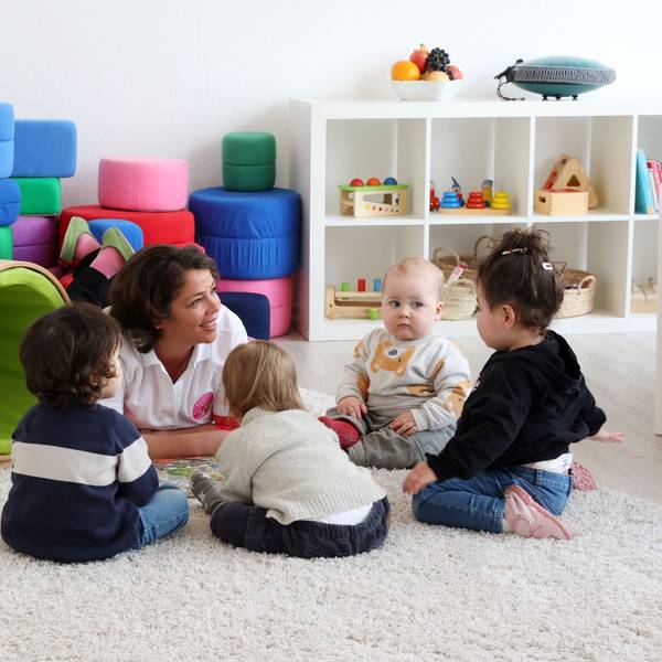 Eine Tagesmutter liegt auf dem Boden und spricht mit vier Kleinkindern, die auf dem Boden sitzen. Im Hintergrund ist Spielzeug zu sehen.