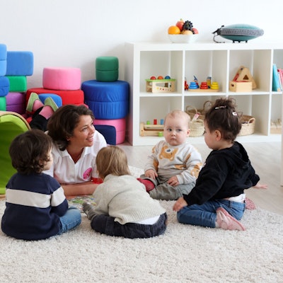 Eine Tagesmutter liegt auf dem Boden und spricht mit vier Kleinkindern, die auf dem Boden sitzen. Im Hintergrund ist Spielzeug zu sehen.