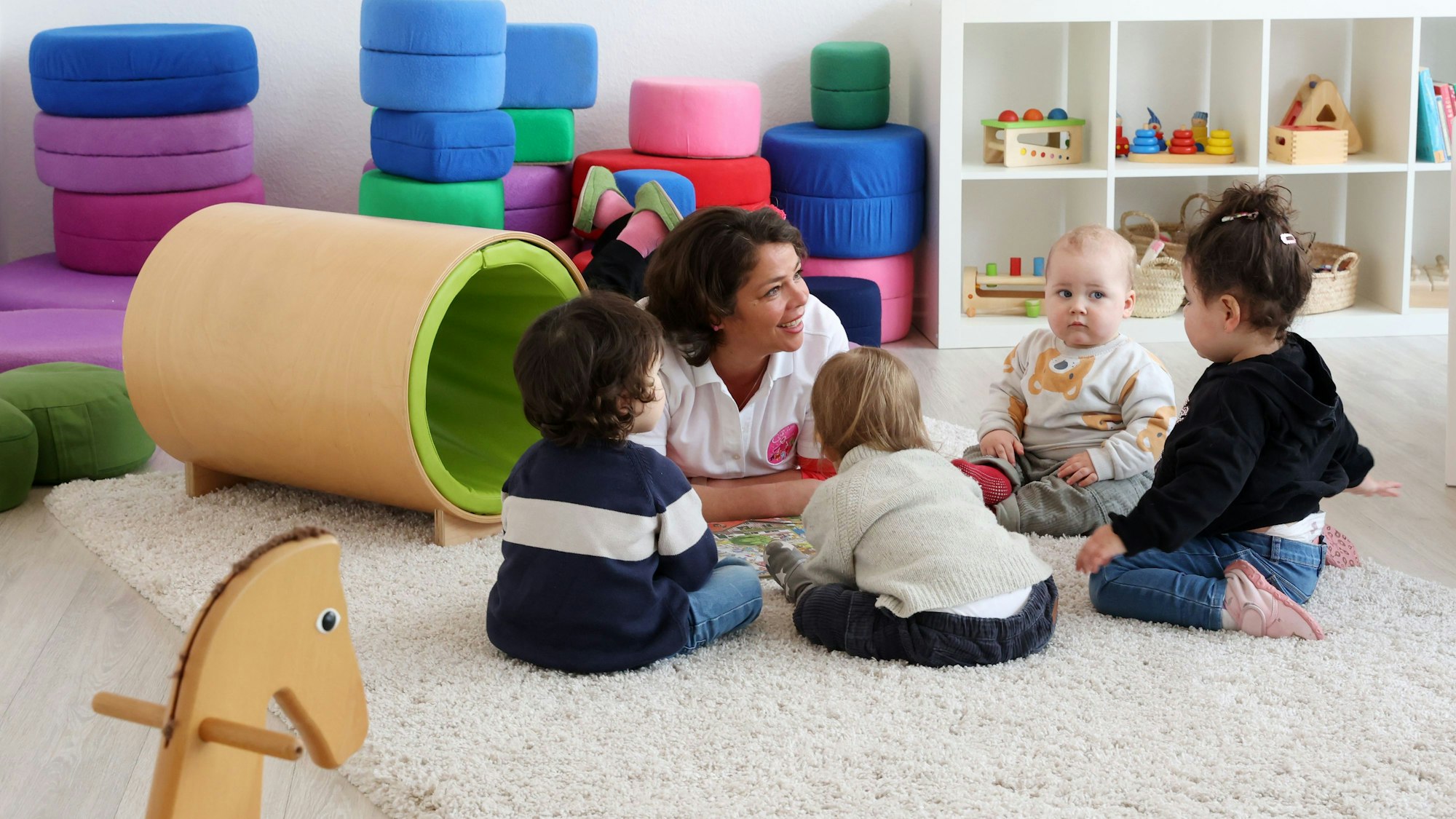 Eine Tagesmutter liegt auf dem Boden und spricht mit vier Kleinkindern, die auf dem Boden sitzen. Im Hintergrund ist Spielzeug zu sehen.