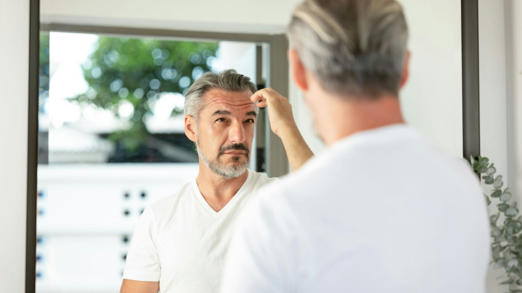 Ein Mann in weißem T-Shirt schaut in einen Spiegel und fährt sich mit der rechten Hand durch seine grauen Haare