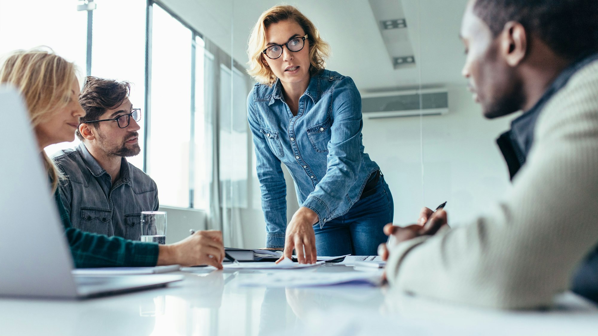 Eine Frau steht an einem Konferenztisch. Um sie herum sitzen mehrere Mitarbeitende.