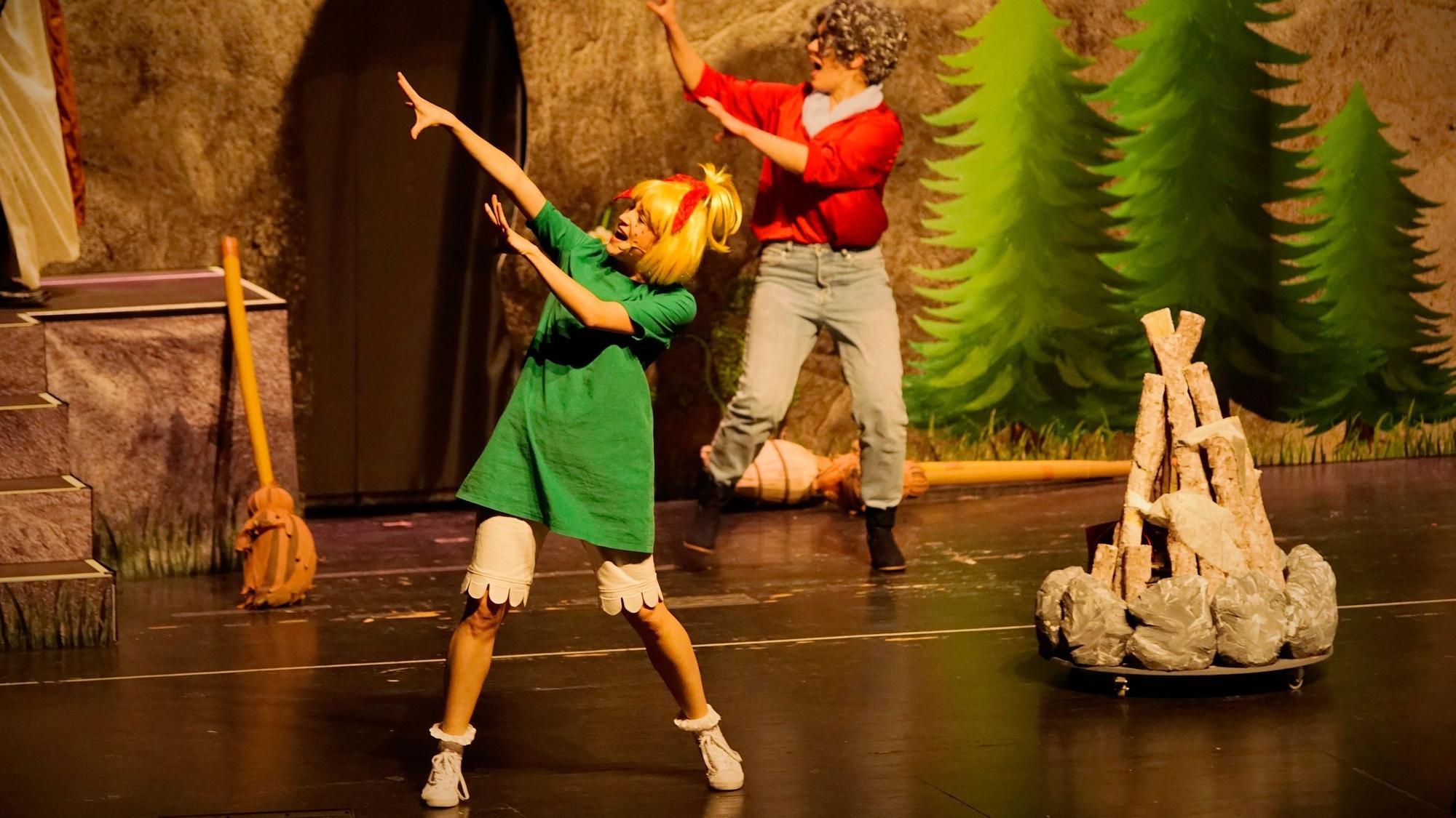 Szene aus dem neuen Bibi-Blocksberg-Musical „Alles wie verhext!“. Frau mit blonder Perücke und grünem T-Shirt-Kleid tanzt vor einem aufgestapelten Lagerfeuer, hinter ihr tanzt eine Frau mit grauer Perücke.