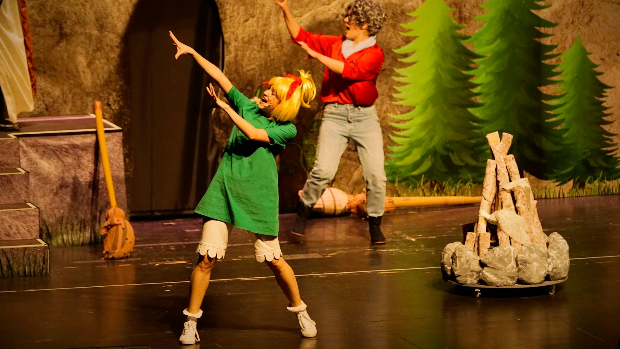 Szene aus dem neuen Bibi-Blocksberg-Musical „Alles wie verhext!“. Frau mit blonder Perücke und grünem T-Shirt-Kleid tanzt vor einem aufgestapelten Lagerfeuer, hinter ihr tanzt eine Frau mit grauer Perücke.