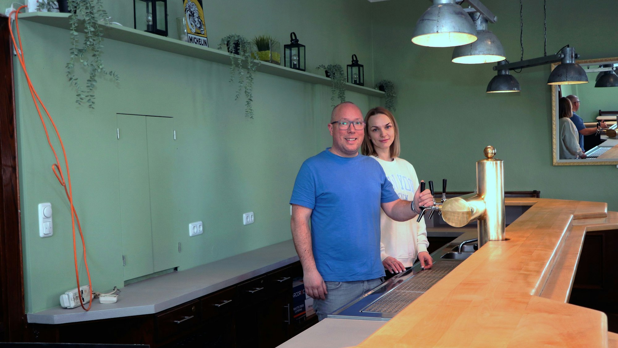 Ein Mann im blauen T-Shirt und eine Frau im weißen Sweatshirt stehen am Zapfhahn hinter einer Theke. Im Hintergrund eine etwas nackt wirkende Wand.