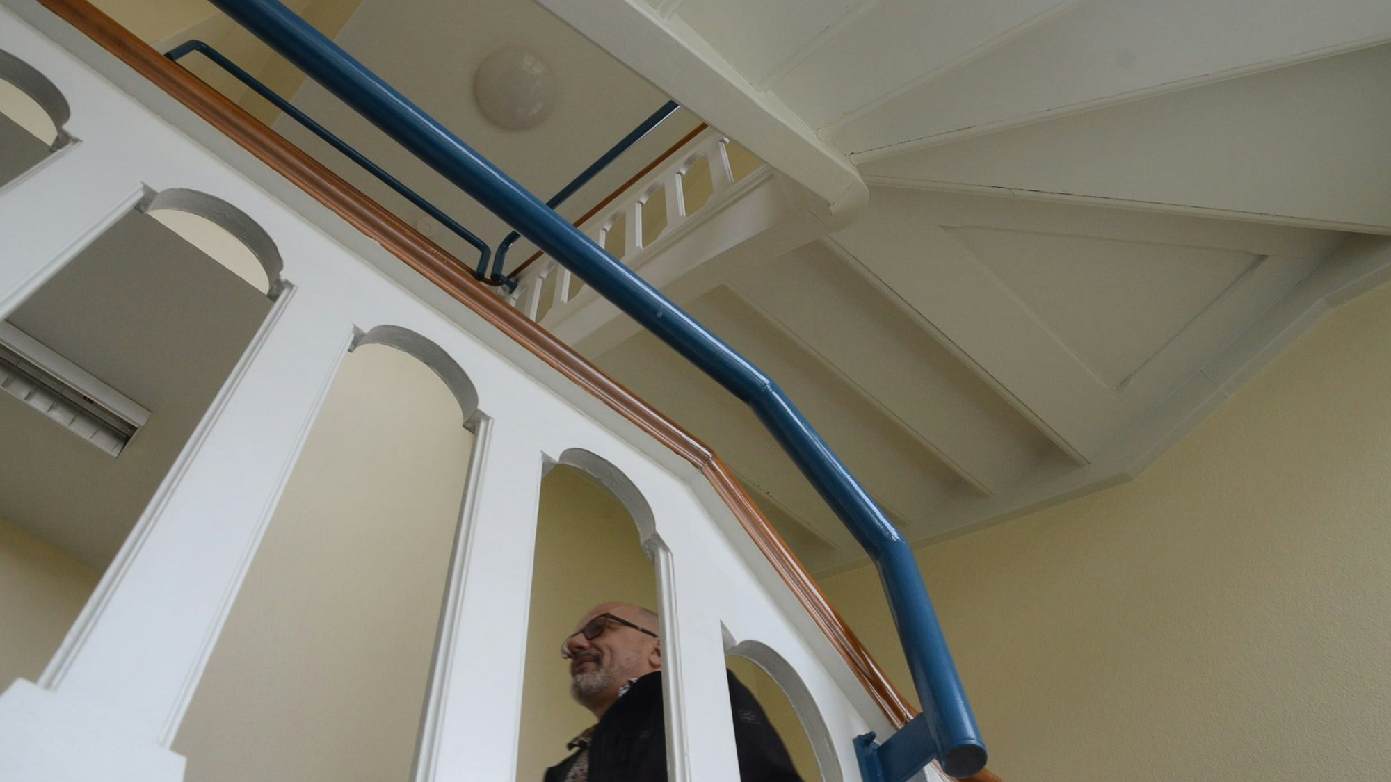 Blick nach oben in einem Treppenhaus. Stufen und Geländer sind weiß lackiert; es gibt einen blauen Handlauf.