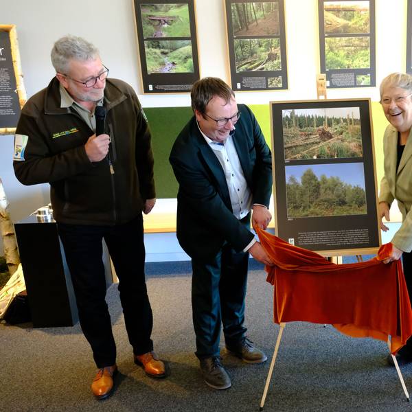 Eröffnung der Sonderausstellung Wilde Bilder. Oliver Krischer und Bärbel Höhn stehen vor einer Staffelei, auf der zwei Fotos präsentiert werden, sie halten ein rotes Tuch. Daneben steht Michael Lammertz mit einem Mikrofon.