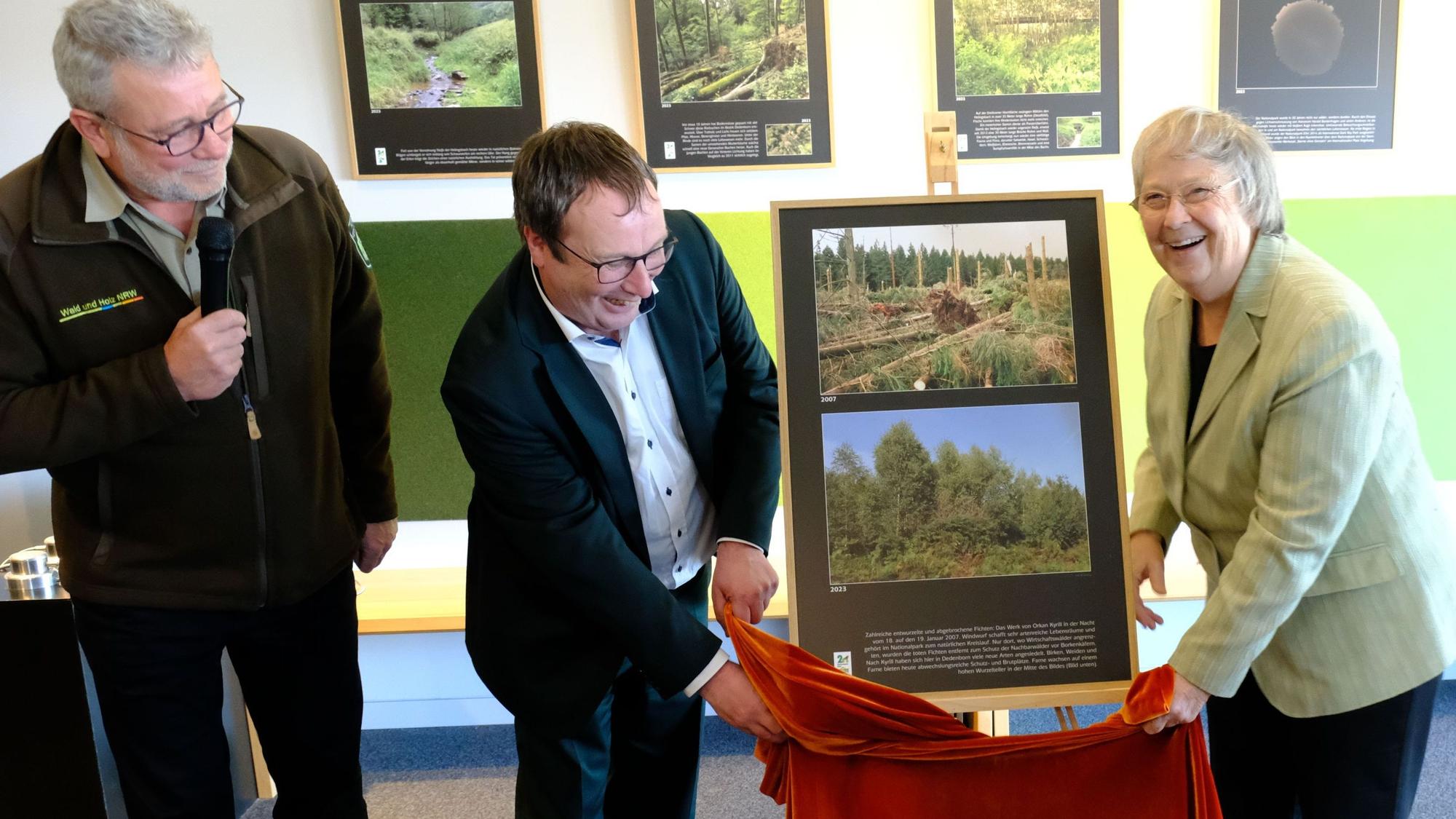 Eröffnung der Sonderausstellung Wilde Bilder. Oliver Krischer und Bärbel Höhn stehen vor einer Staffelei, auf der zwei Fotos präsentiert werden, sie halten ein rotes Tuch. Daneben steht Michael Lammertz mit einem Mikrofon.