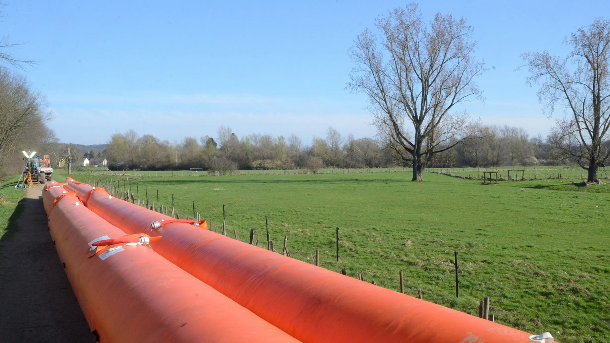 Zwei leuchtend orange Kunststoffschläuche liegen auf einem Deich oberhalb der Flussaue.