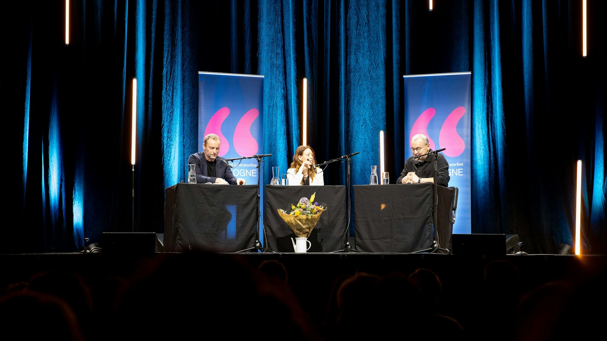 Mark Waschke, Lavinia Wilson und Knut Elstermann sitzen an drei Tischen auf der Bühne. Im Hintergrund ein blauer Vorhang und einige längliche LED-Röhren, von denen manche zu schweben scheinen-