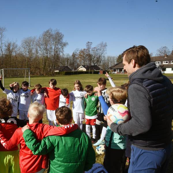 Eine Gruppe Kinder und zwei Trainer stehen in einem Kreis auf einem Fußballplatz zusammen.