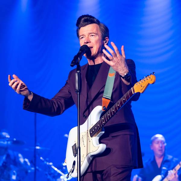 Rick Astley gestikuliert am Mikrofon auf der Bühne des Kölner Palladiums. Er trägt einen auberginefarbenen Zweireiher und hat eine weiße E-Gitarre umgeschnallt.