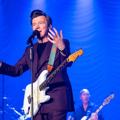 Rick Astley gestikuliert am Mikrofon auf der Bühne des Kölner Palladiums. Er trägt einen auberginefarbenen Zweireiher und hat eine weiße E-Gitarre umgeschnallt.