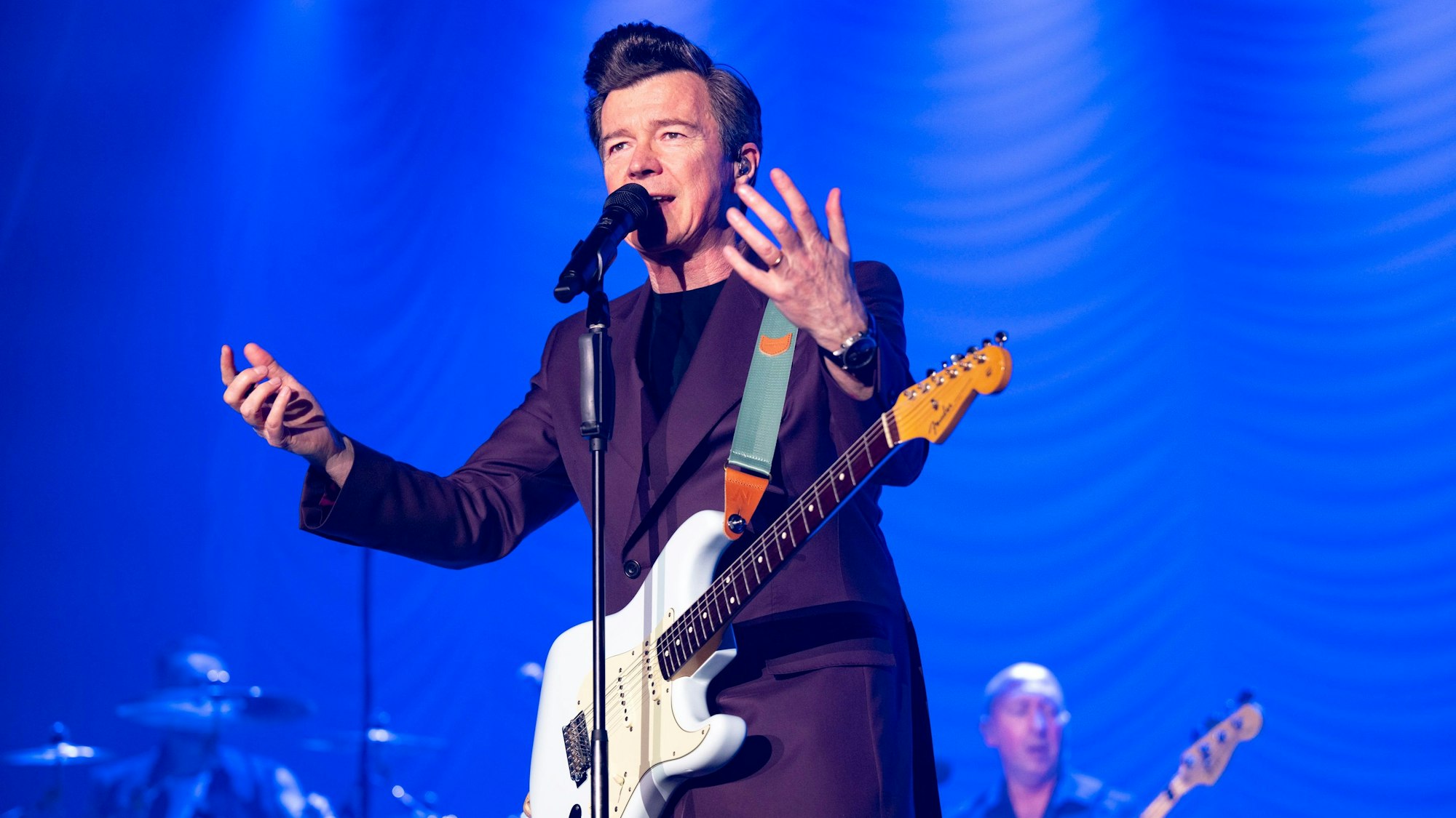 Rick Astley gestikuliert am Mikrofon auf der Bühne des Kölner Palladiums. Er trägt einen auberginefarbenen Zweireiher und hat eine weiße E-Gitarre umgeschnallt.