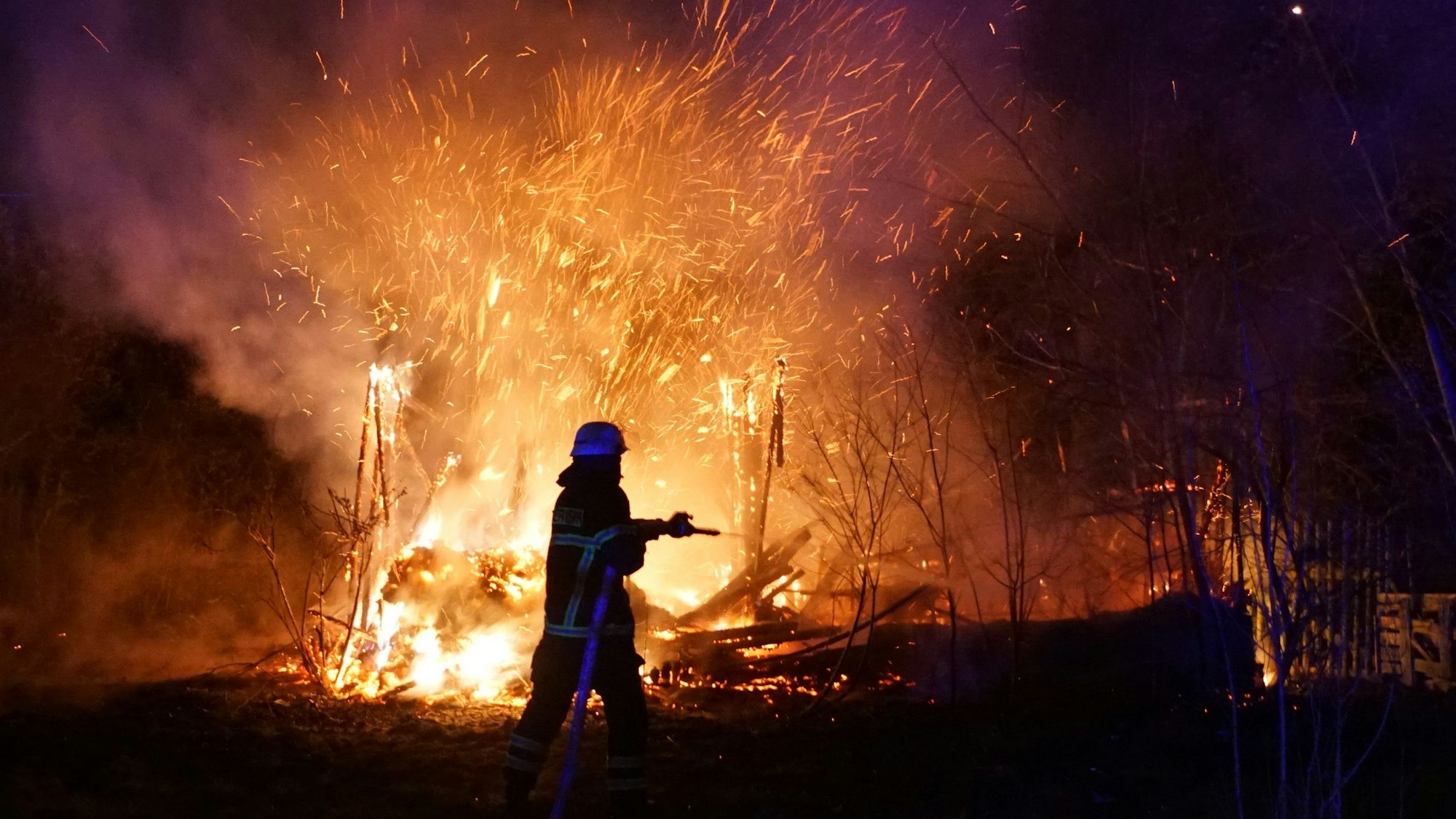 Eine Einsatzkraft der Feuerwehr steht mit einem Strahlrohr vor einem brennenden Schuppen. Funken schlagen in den Nachthimmel.