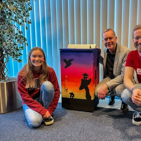 Eine Jugendliche und zwei Männer knien neben einem bemalten Stromkasten.