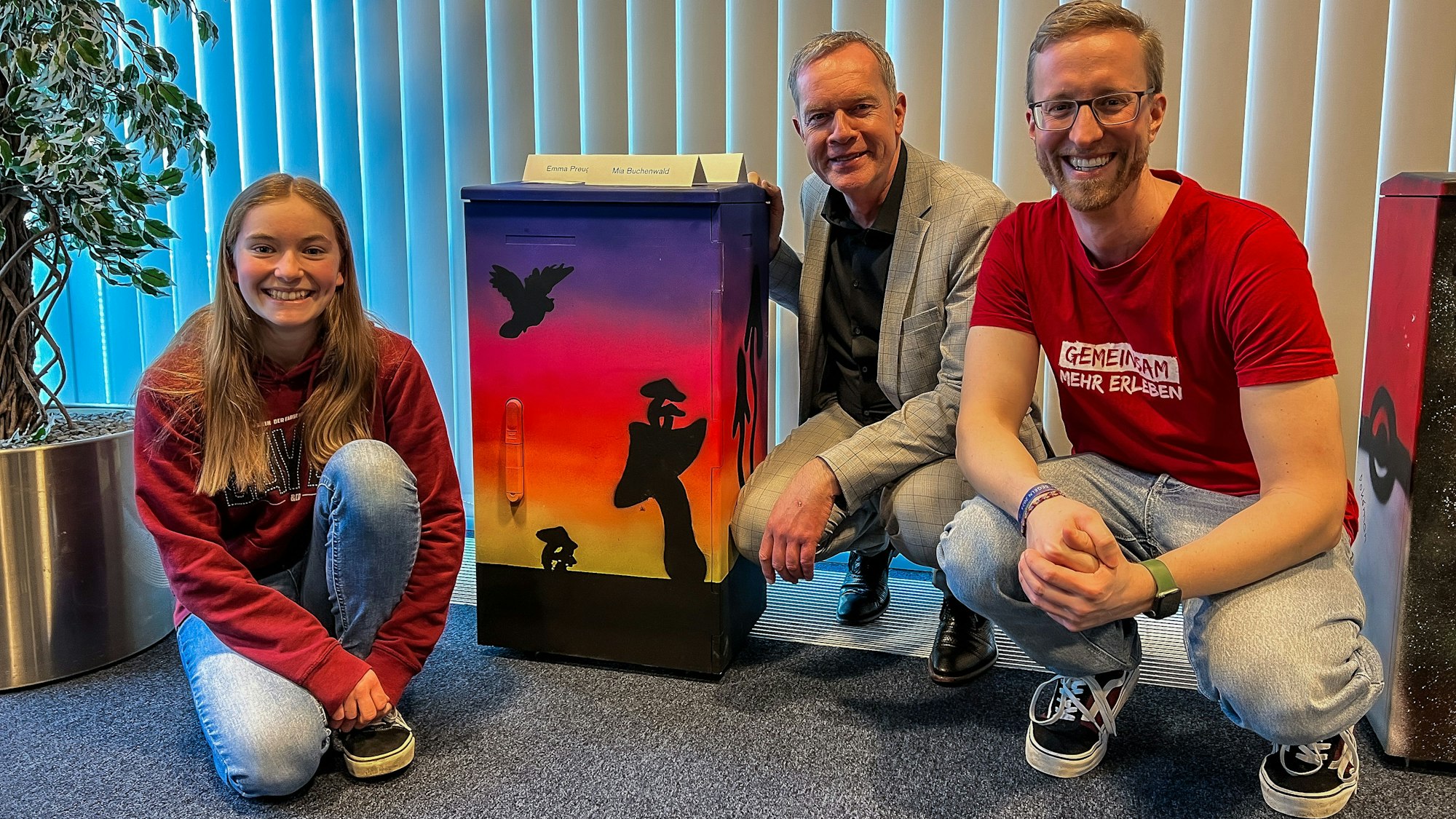 Eine Jugendliche und zwei Männer knien neben einem bemalten Stromkasten.