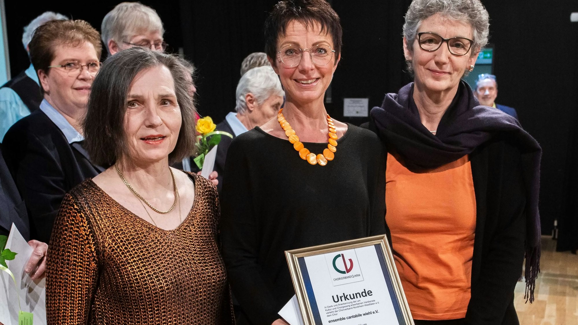 Drei Frauen stehen nebeneinander. Eine Frau hält eine Urkunde in der Hand.