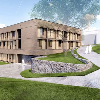 Ein langgestreckter Bau aus Holz ist umgeben von Fußwegen und einer Grünanlage.