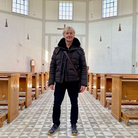 Ein blonder Mann mit Daunenjacke und Umhängetasche steht in einem hellen, großen Raum mit Kirchenbänken und gemustertem Fußboden.