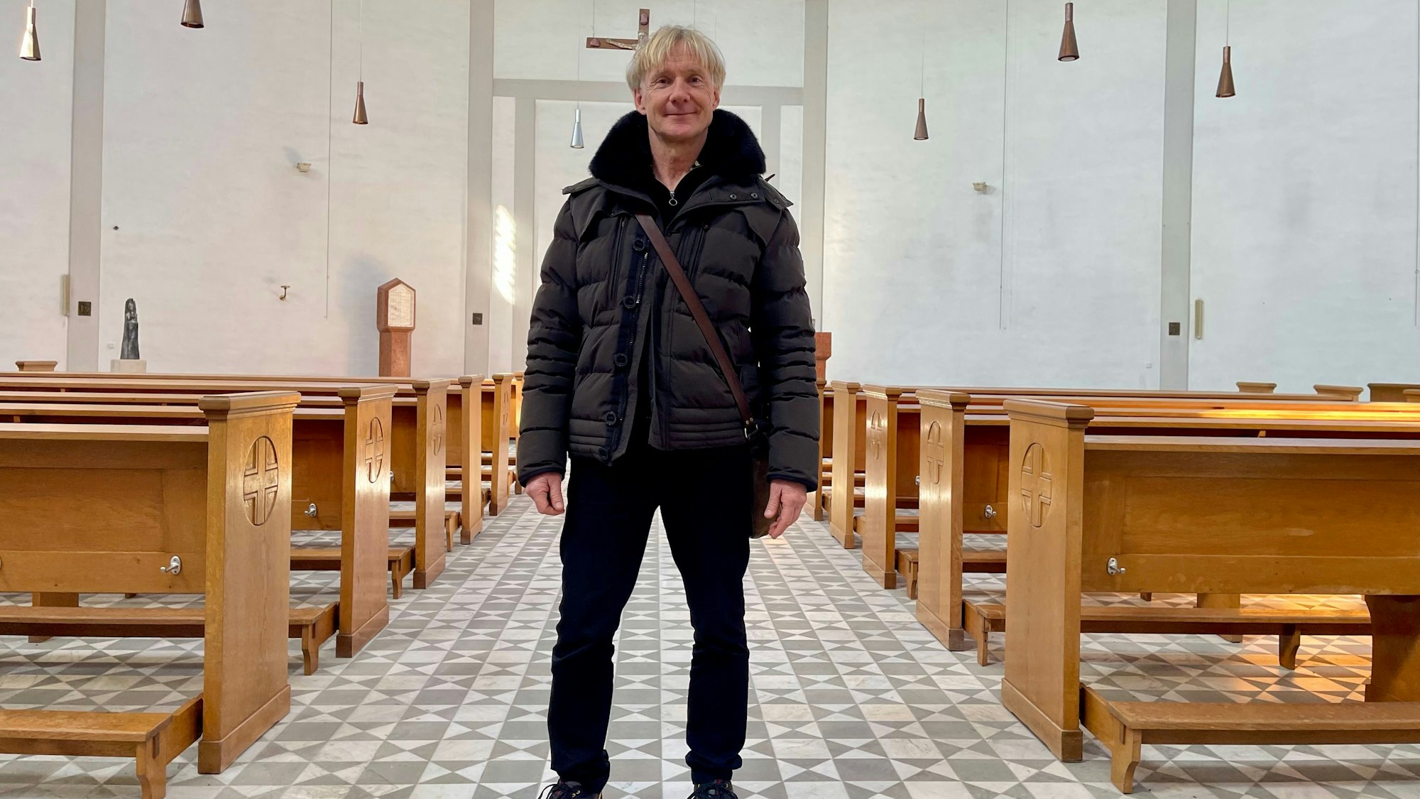 Ein blonder Mann mit Daunenjacke und Umhängetasche steht in einem hellen, großen Raum mit Kirchenbänken und gemustertem Fußboden.