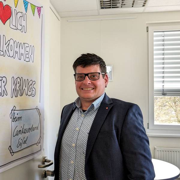 Oliver Krings steht an der Tür seines Büros, an dem ein Plakat mit der Aufschrift „Herzlich willkommen Oliver Krings“ zu sehen ist.