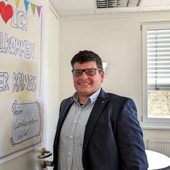 Oliver Krings steht an der Tür seines Büros, an dem ein Plakat mit der Aufschrift „Herzlich willkommen Oliver Krings“ zu sehen ist.