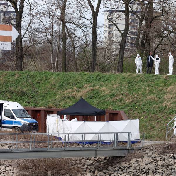 Polizisten der Spurensicherung und ein Staatsanwalt stehen auf einer Böschung im Mülheimer Hafen und schauen auf einen schmalen Weg.