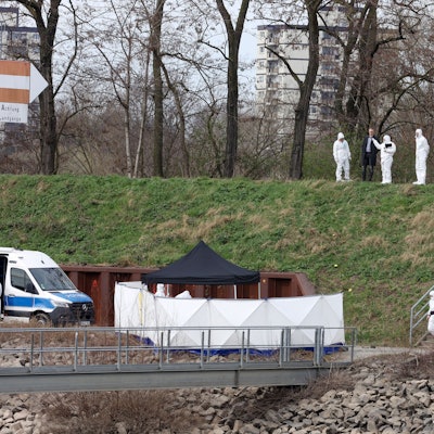 Polizisten der Spurensicherung und ein Staatsanwalt stehen auf einer Böschung im Mülheimer Hafen und schauen auf einen schmalen Weg.