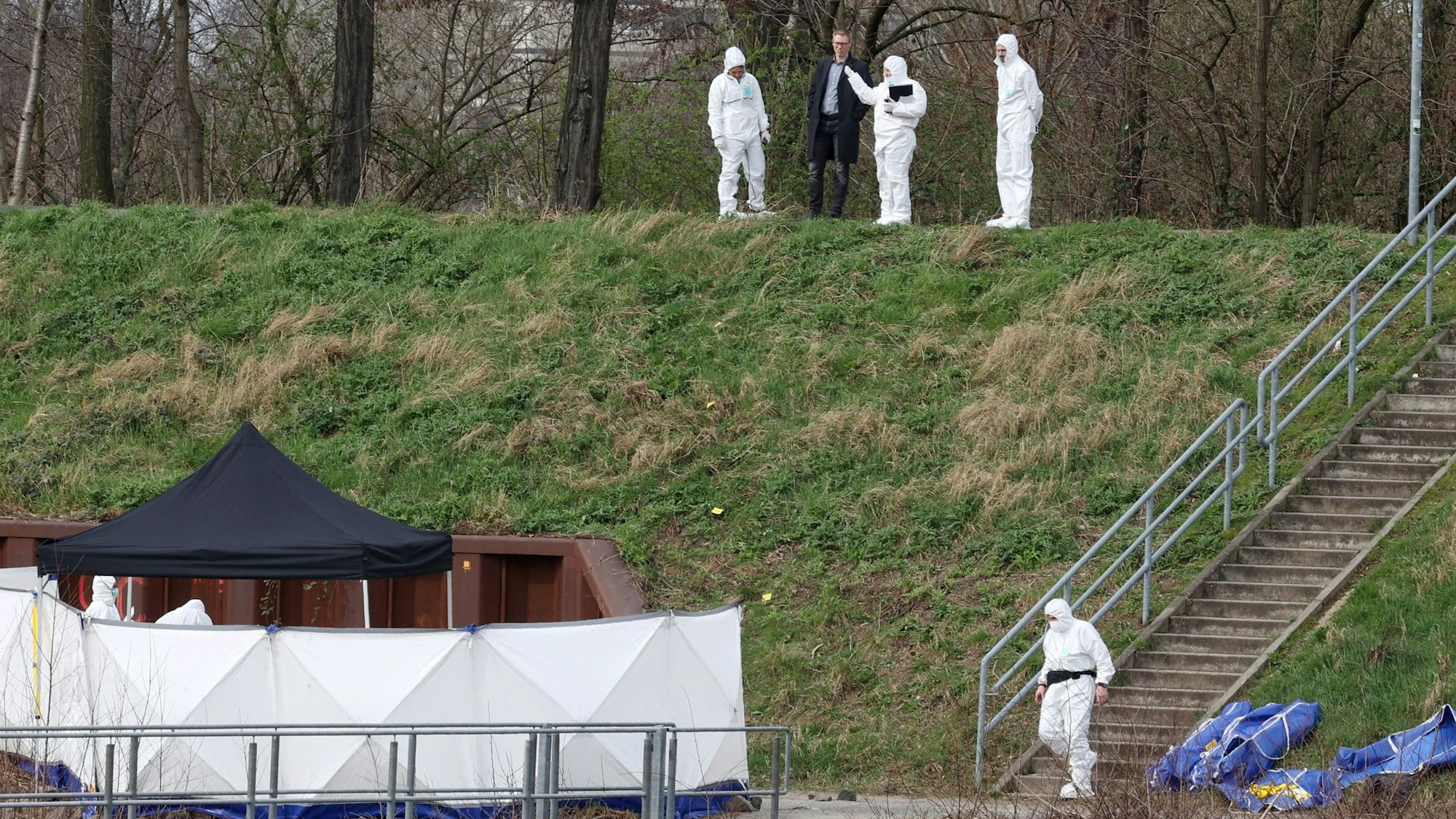Polizisten der Spurensicherung und ein Staatsanwalt stehen auf einer Böschung im Mülheimer Hafen und schauen auf einen schmalen Weg.