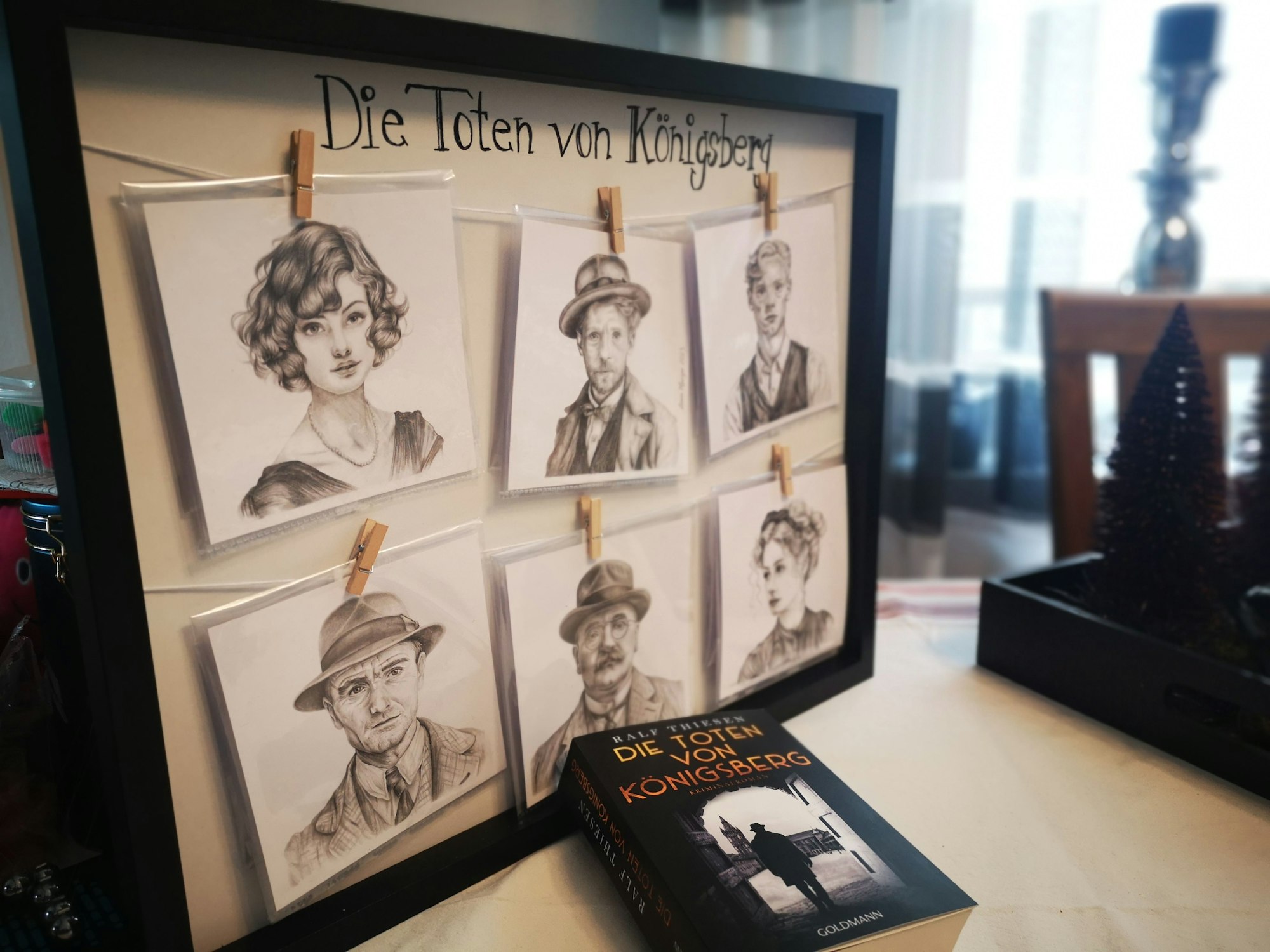 Ein Rahmen mit gezeichneten Porträts steht auf einem Tisch. Sechs Personen sind in schwarz-weiß abgebildet. Auf dem Rahmen steht die Überschrift „Die Toten von Königsberg“