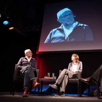 Die drei Gäste sitzen auf gepolsterten Stühlen auf der Bühne. Hinter ihnen hängt eine große Leinwand, auf der das Bühnengeschehen projiziert wird.