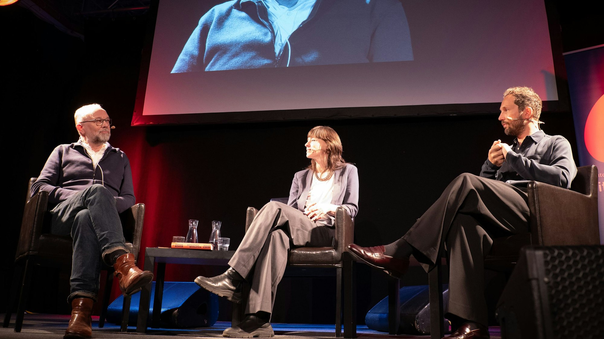 Die drei Gäste sitzen auf gepolsterten Stühlen auf der Bühne. Hinter ihnen hängt eine große Leinwand, auf der das Bühnengeschehen projiziert wird.