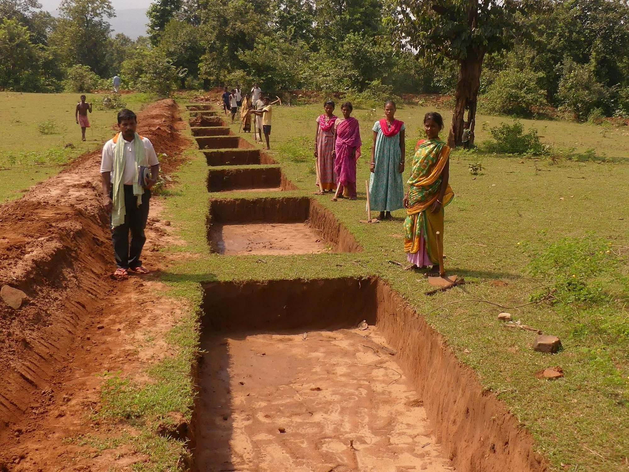Entlang einer landwirtschaftlichen Fläche sind mehrere rechteckige Gruben ausgehoben worden. Einige Arbeiterinnen und Arbeiten schauen in die Kamera.