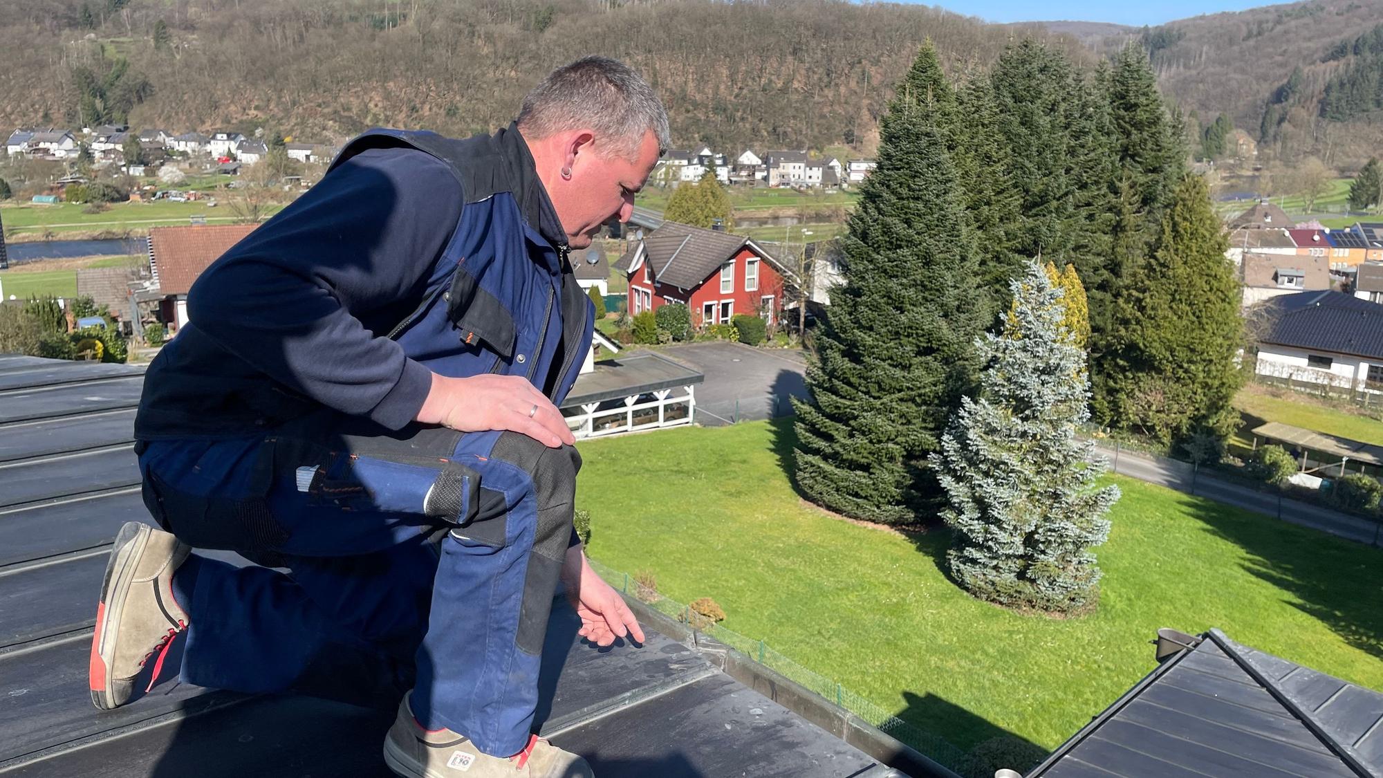 Ein Mann in einem blauen Arbeitsoverall kniet auf einen Dach.
