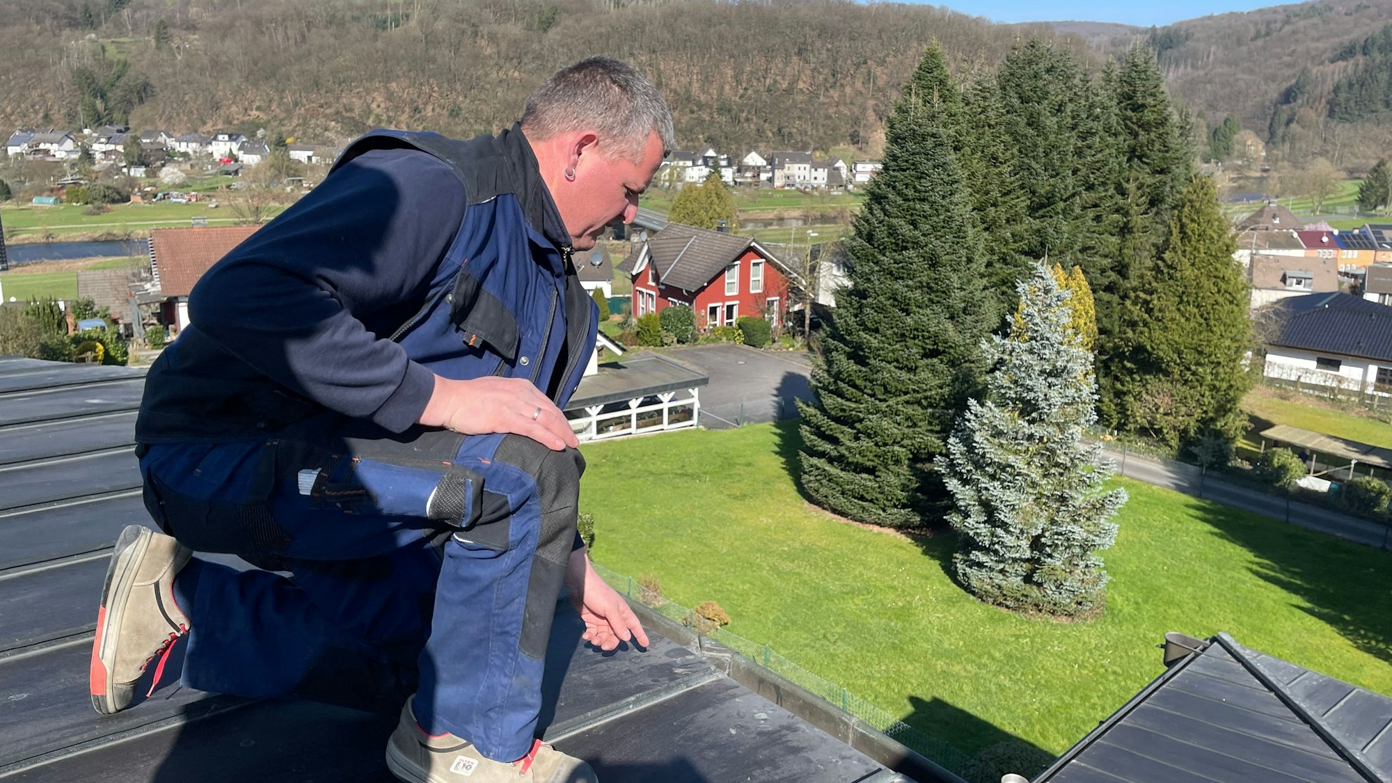 Ein Mann in einem blauen Arbeitsoverall kniet auf einen Dach.