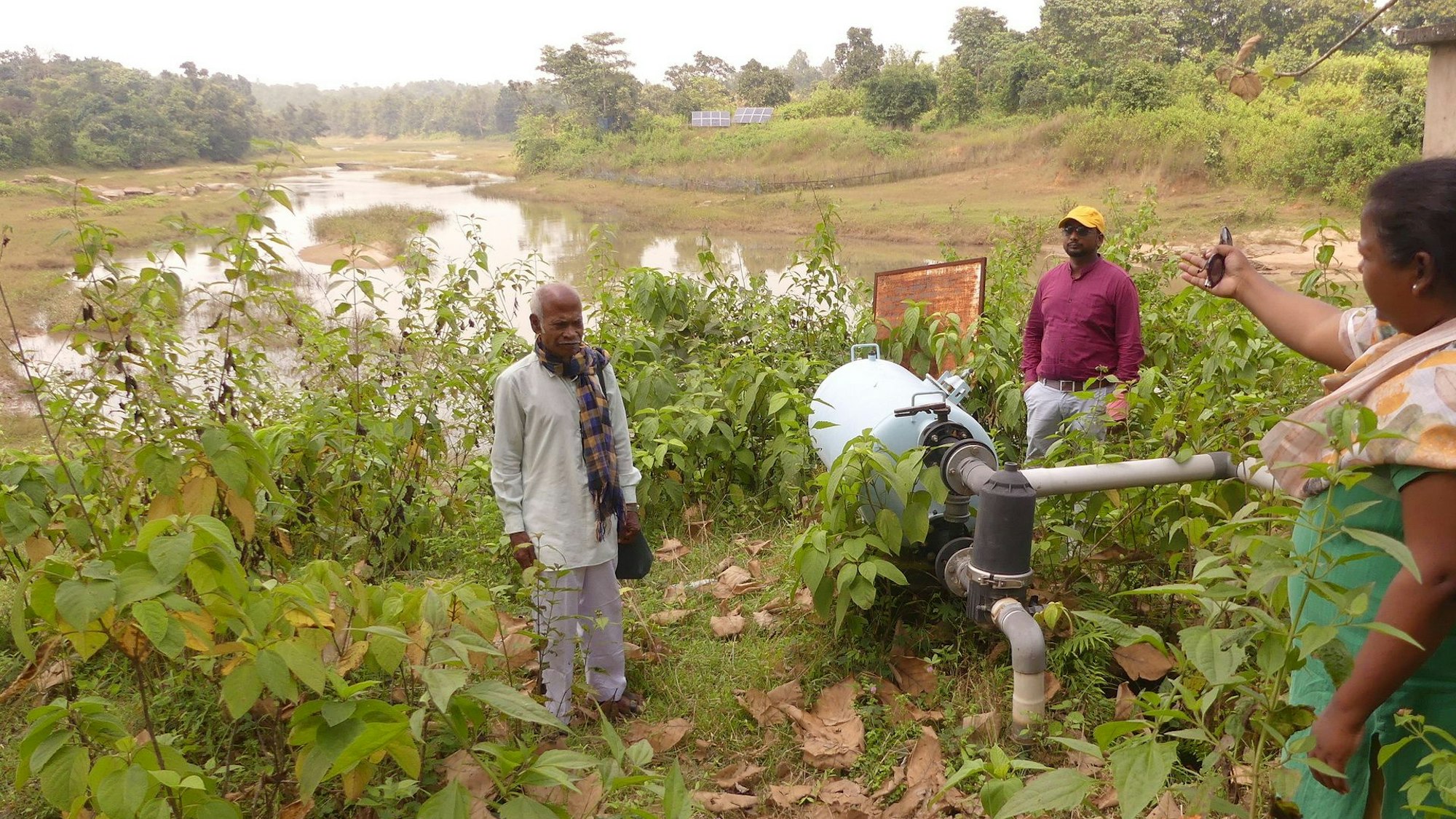 Eine Frau deutet mit ausgestreckter Hand auf einen mit Wasser gefüllten Teich im Hintergrund. Neben einer Pumpe stehen zwei weitere Männer.