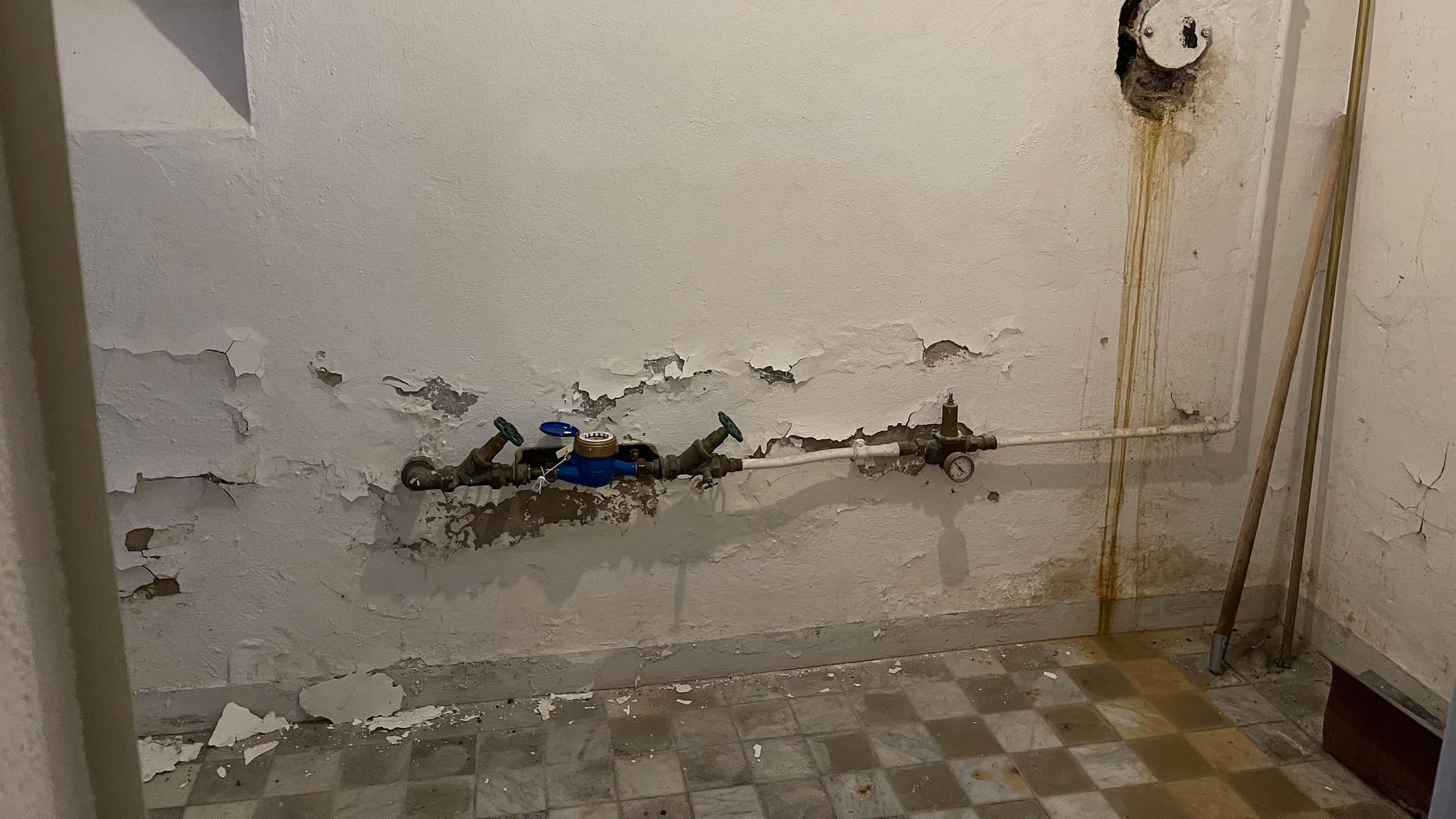 Von einer weiß verputzten Wand mit Rohren bröckelt Putz, der karierte Steinboden ist mit bräunlichem Wasser bedeckt.