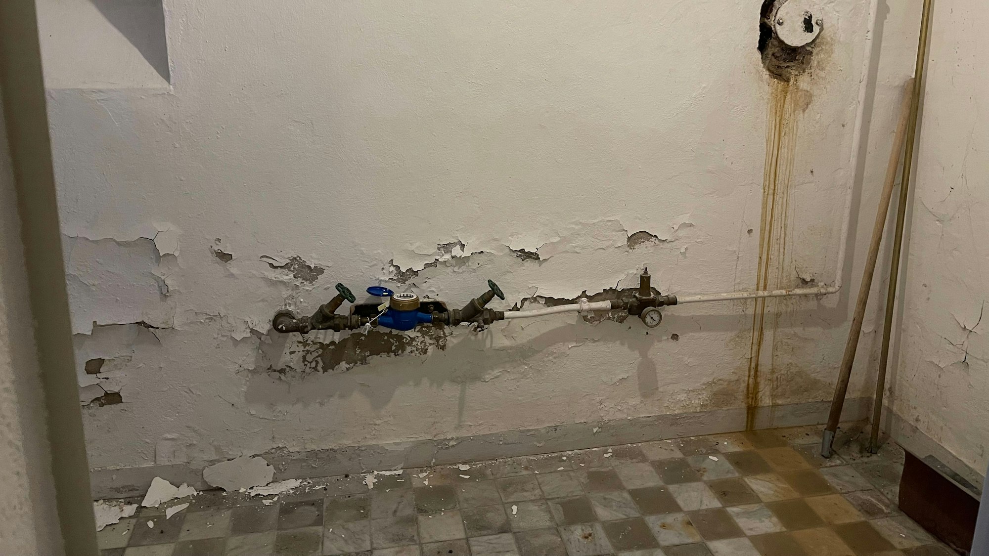 Von einer weiß verputzten Wand mit Rohren bröckelt Putz, der karierte Steinboden ist mit bräunlichem Wasser bedeckt.