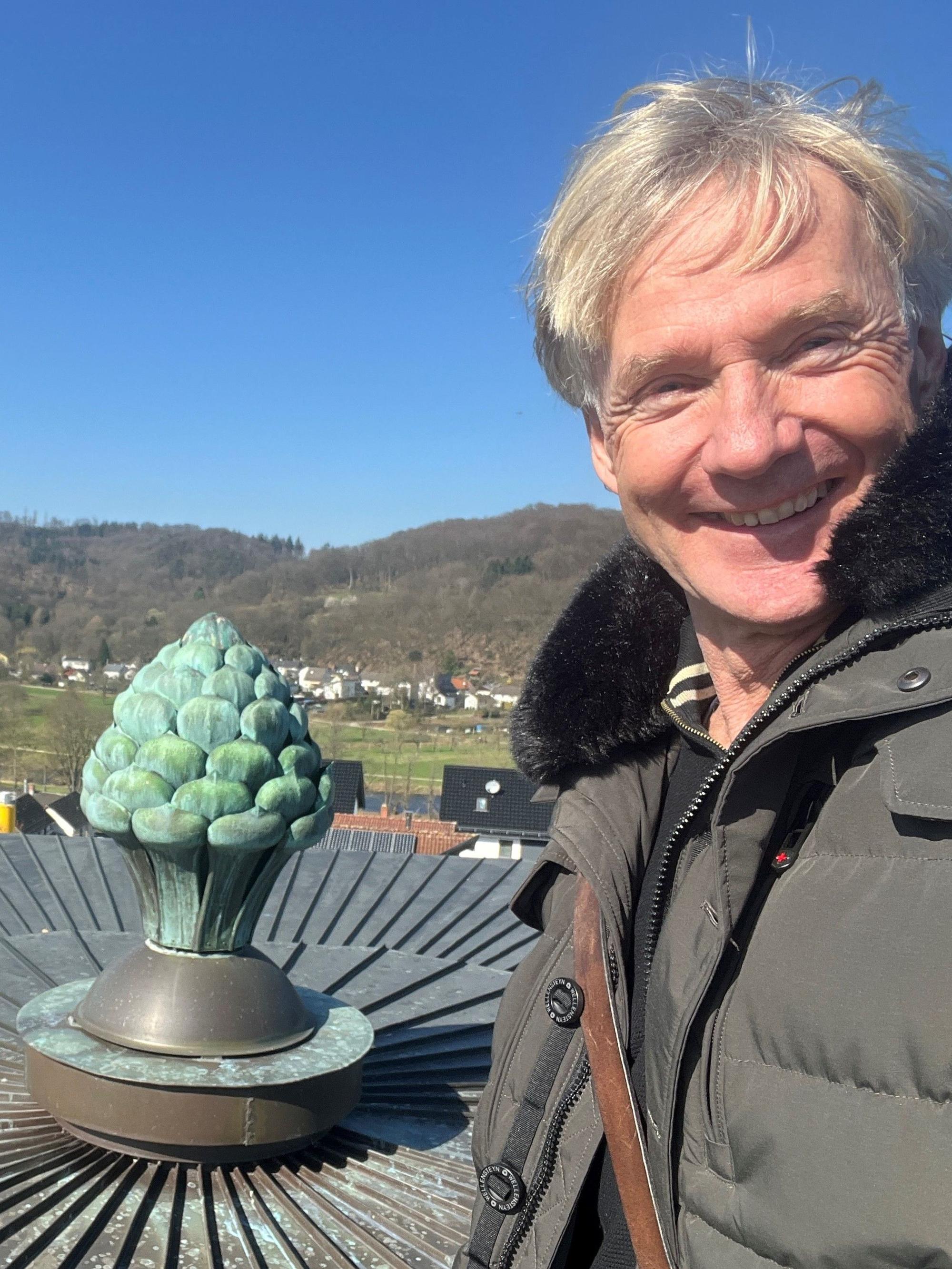 Ein blonder Mann lacht in die Kamera, neben ihm ein großer Pinienzapfen aus Kupfer.