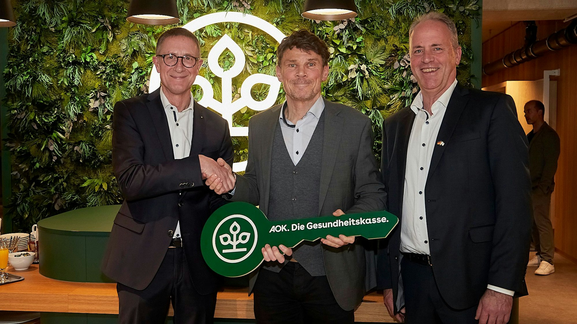 Drei Männer stehen vor einer mit grünen Pflanzen bewachsenen Wand. Zwei geben sich die Hand, einer hält einen großen Schlüssel mit dem Logo und Schriftzug der Krankenkasse AOK.