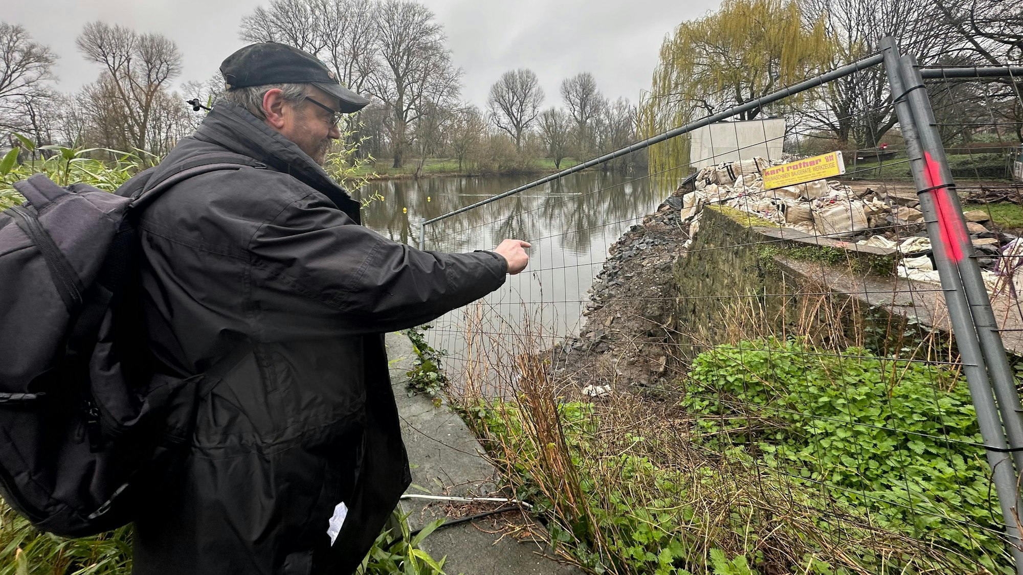 Ein Mann mit Kappe und zeigt auf eine Stelle in einer Mauer an einem See.