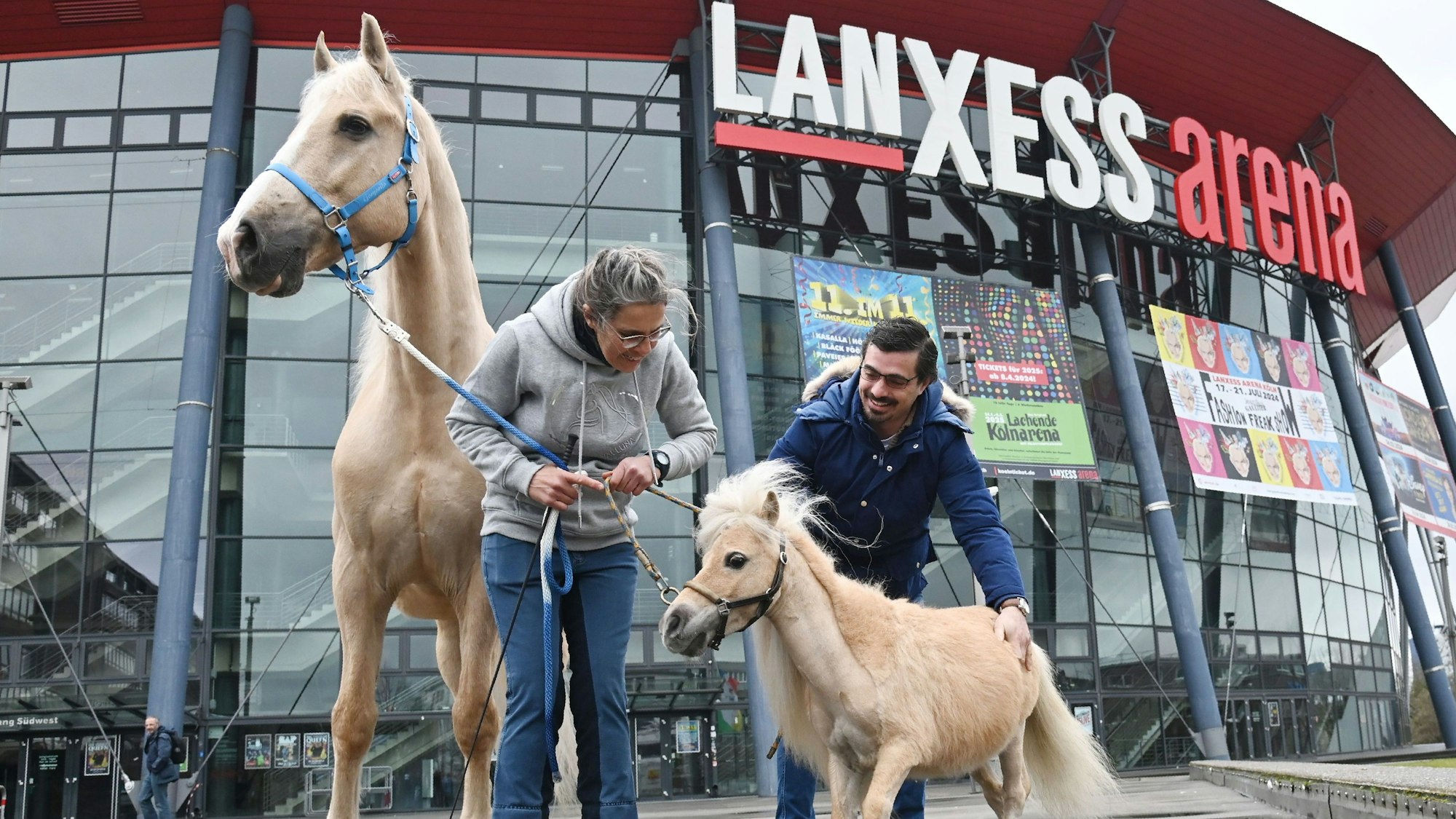Auf dem Foto sind eine Frau und ein Mann sowie zwei Pferde vor der Lanxess Arena zusehen.