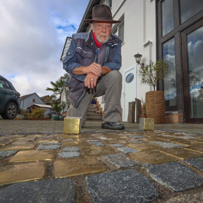 Künstler Gunter Demnig arbeitet goldene Stolpersteine in das Pflaster einer Straße ein.