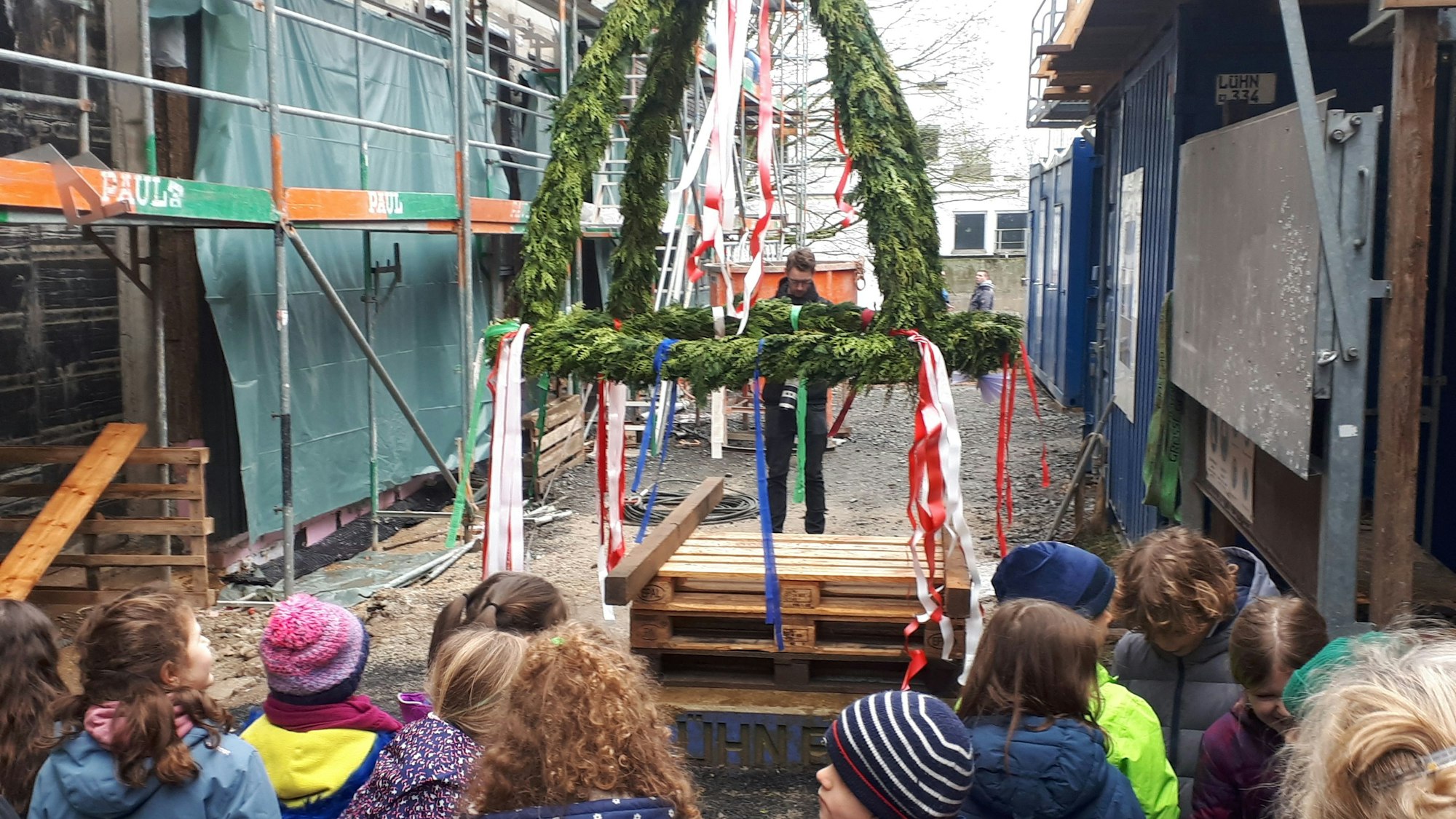 Kinder stehen auf einer Baustelle vor einem festlich geschmückten Richtkranz.