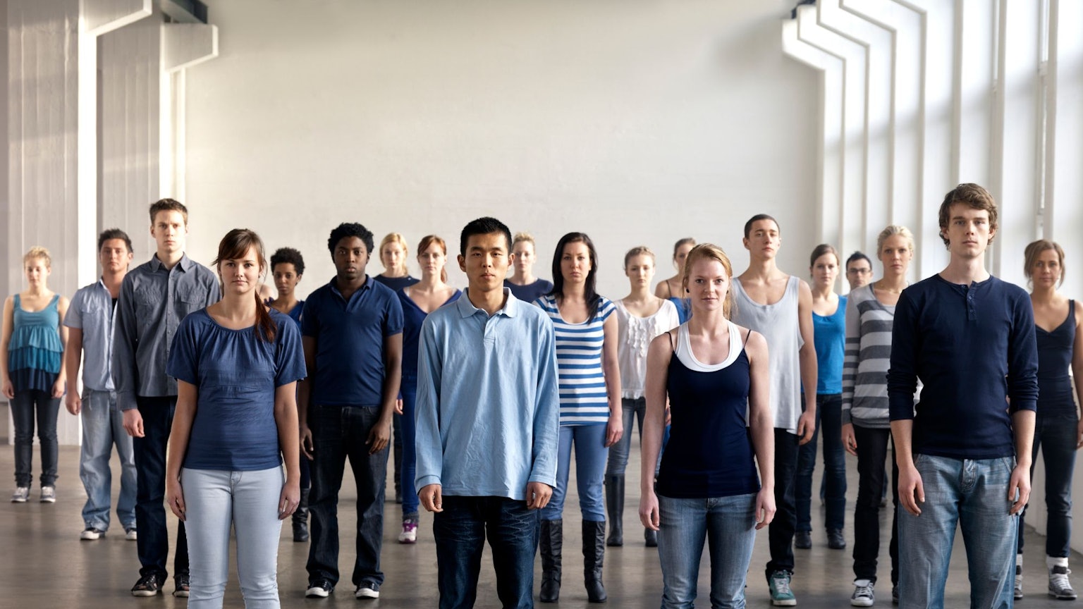 Eine Gruppe junger Menschen verschiedenster Herkunft steht in einem Raum, sie alle blicken in die Kamera.