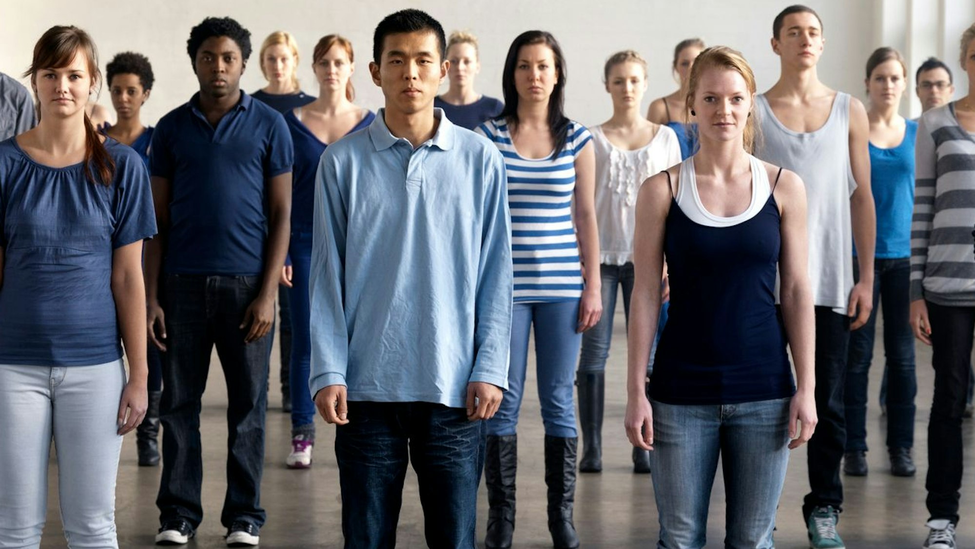 Eine Gruppe junger Menschen verschiedenster Herkunft steht in einem Raum, sie alle blicken in die Kamera.