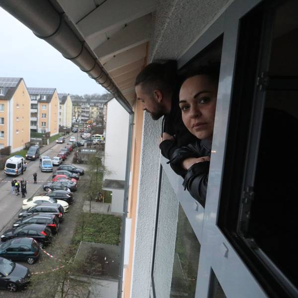 Eine Frau schaut aus einem Fenster auf den abgesperrten Tatort vor einem Wohnhaus in Vingst.