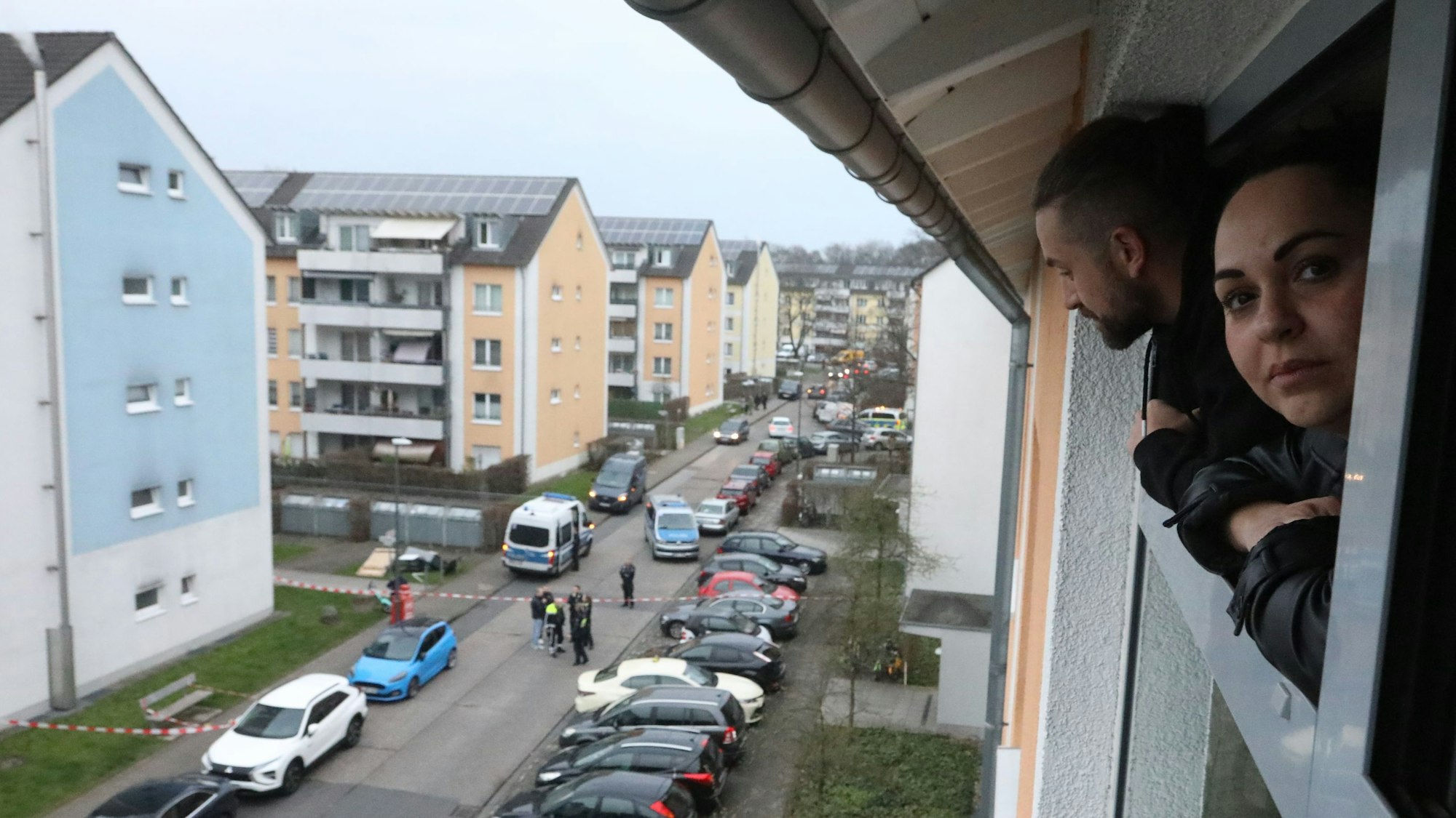Eine Frau schaut aus einem Fenster auf den abgesperrten Tatort vor einem Wohnhaus in Vingst.