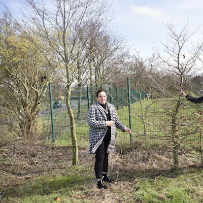 Juliane B. Vetter und Julia Drittler stehen mit einem Baum in der Hand vor einem grünen Zaun.
