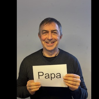 Der Vater hält einen Zettel vor der Brust mit dem Schriftzug „Papa“.